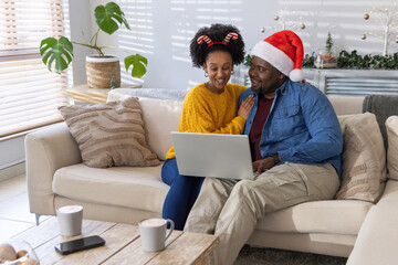 African American couple sitting on sofa wearing Santa hat and candy-cane headband sharing laptop