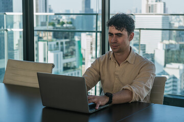 Fototapeta premium Man focusing on his laptop in a contemporary office setting with a city skyline view, symbolizing remote work and urban professionalism