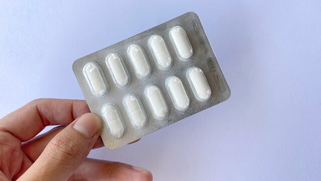 A hand holding a blister pack containing white medication tablets against a clean white background. Showing pharmaceutical product details and illustrating the concept of medical care.
