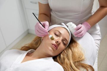 Naklejka premium Cosmetologist applying a mask from clay to the face of a beautiful woman. Spa treatment and face care in the beauty salon