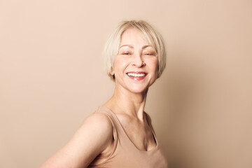 Fototapeta premium Smiling mature woman with short blonde hair wearing beige tank top poses against plain background. Concept of happiness, confidence, natural beauty, and positive aging.
