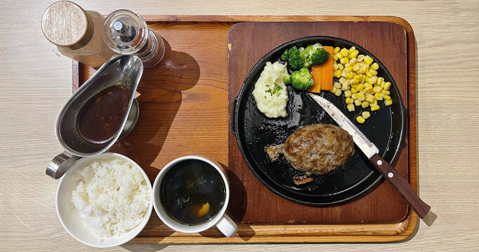 Top view of japanese beef hamburg steak with vegetables, rice, soup, and brown sauce served on wooden tray in restaurant setting, balanced meal concept.