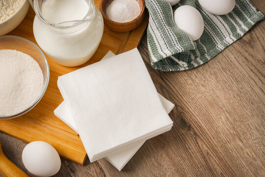 A stack of square white paper napkins lies on a wooden board next to a glass jug of milk, a bowl of sugar, and a towel with eggs. Copy space. Photo.
