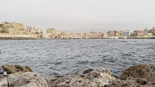 Slow reveal of Chania's Old Venetian Port, the main tourist area, with warm golden hour glow at sunset and hypnotising waves of the Cretan Sea [Chania, Crete, Greece]