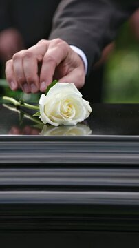 Man gently placing a single white rose on a black coffin lid during an outdoor funeral ceremony, vertical shot