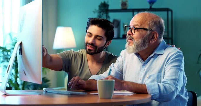Indian young son teaching senior father how to use desktop computer at home in friendly way, elderly dad learning digital technology with smile while browsing internet together in modern living room