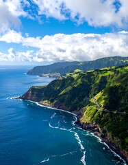 Aerial view of a rugged coastline meeting a vibrant green hillside under a partly cloudy, bright blue sky. Waves crash below