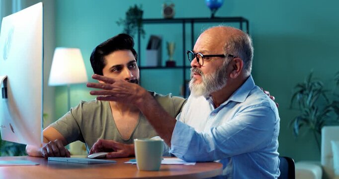 Indian young son teaching senior father how to use desktop computer at home in friendly way, elderly dad learning digital technology with smile while browsing internet together in modern living room