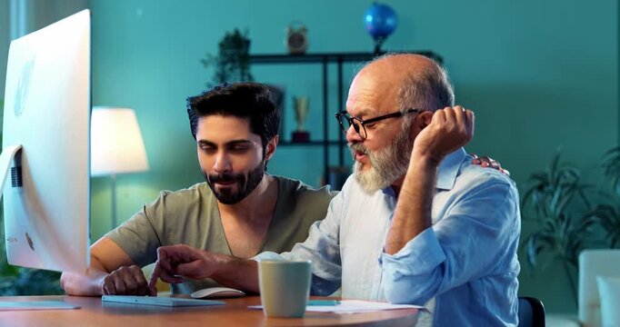 Indian young son teaching senior father how to use desktop computer at home in friendly way, elderly dad learning digital technology with smile while browsing internet together in modern living room