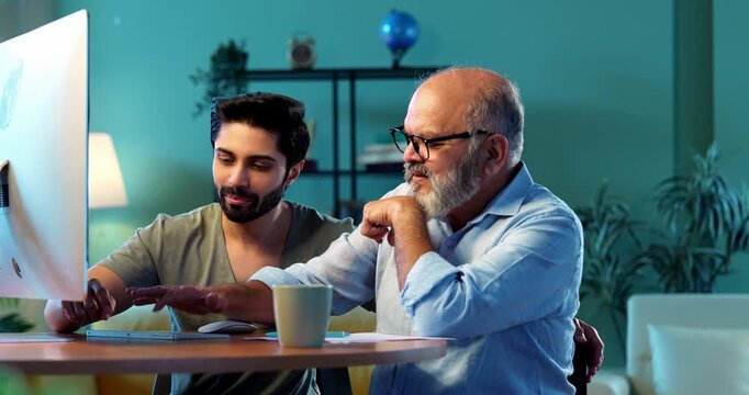 Indian young son teaching senior father how to use desktop computer at home in friendly way, elderly dad learning digital technology with smile while browsing internet together in modern living room