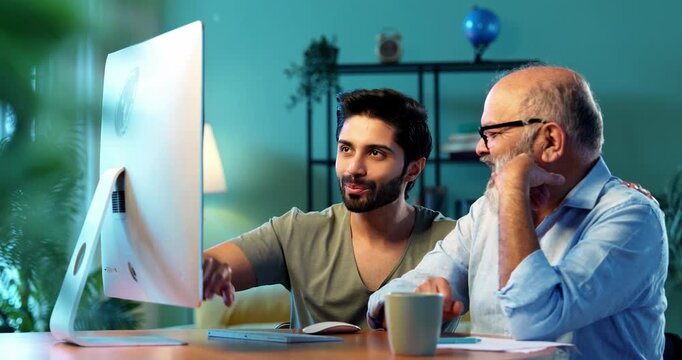 Indian young son teaching senior father how to use desktop computer at home in friendly way, elderly dad learning digital technology with smile while browsing internet together in modern living room
