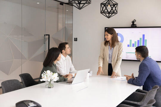 Middle-aged female boss, corporate trainer or executive provide explanations for staff members, company client gathered together in conference room with digital screen displaying charts and bar graphs