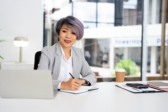 Asian Business woman planning, taking notes and writing in a paper while reading a report, document or paperwork in an office alone at work. One serious corporate professional working at a desk.