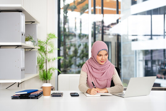 Confident Muslim business woman in hijab, writer  using laptop computer, taking notes, working project in modern office.  financier working at workplace. Successful business.