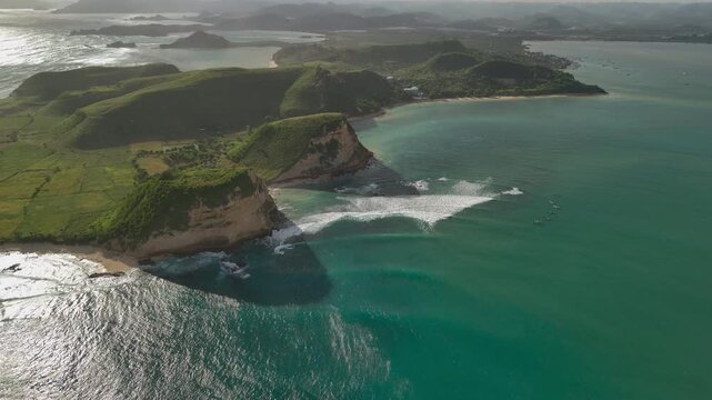 Aerial drone footage of Gerupuk surf bay with reef break white surf lines turquoise water sea cliffs green hills and small boats offshore cinematic coastal seascape in warm evening light 4k