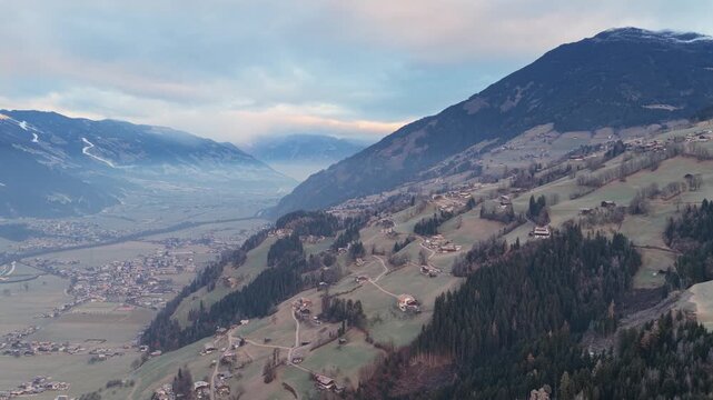 Aerial view of the Kaltenbach valley in Austria during sunrise, with soft light touching alpine meadows and mountain slopes. Morning mist lingers above fields while small houses and winding roads beco