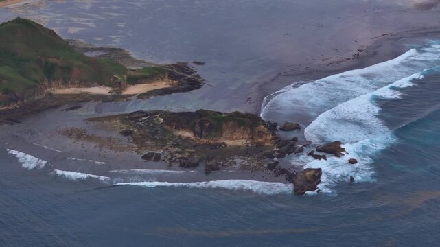 Aerial drone footage of reef break surf wrapping around rocky headland at low tide with seaweed farm grid patterns exposed reef flats whitewater lines and deep blue ocean contrast in 4k