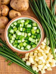 Chopped green onions are neatly arranged in a bowl beside rustic potatoes and chives on a wooden table, showcasing vibrant colors