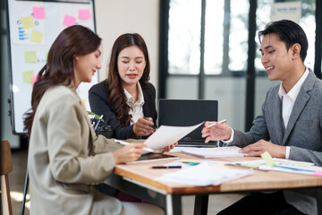Fototapeta premium Group of young Asian professionals engaging in a dynamic brainstorming session. A female leader stands confidently while presenting ideas to her colleagues, showcasing a collaborative, positive
