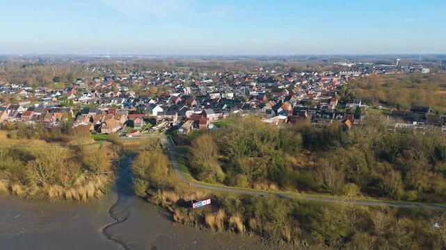 dense belgian riverside town rows brick houses muddy scheldt riverbanks leafless trees wetlands under clear blue sky sint amands belgium wide drone view sint-amands 