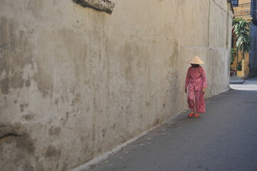 A Vietnamese lady wearing traditional Vietnamese attire with n&oacute;n l&aacute; (conical hat) along an empty narrow street in historical city of Hoi An, Vietnam. 