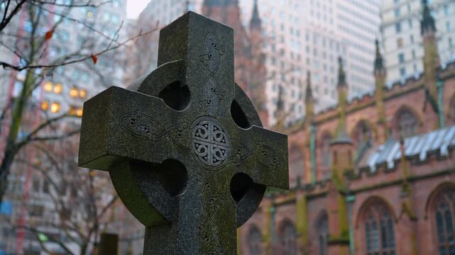 Close up of stone celtic cross headstone in cemetery. Detailed view of an engraved stone cross with a historic church and city buildings in a blurred background.