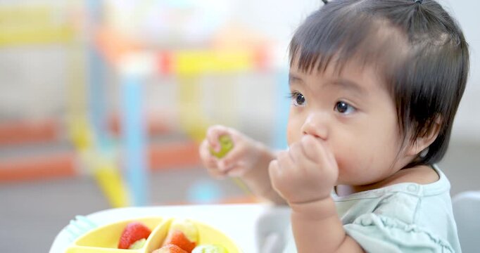 Asian toddler girl sitting on feeding chair holds green grape and looks aside curiously, soft daylight on face, gentle expression, bright background toys, early self feeding moment, child learning