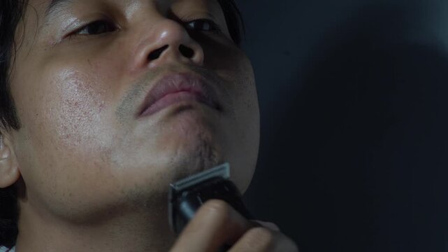 Close-up of a young Southeast Asian man shaving his beard and stubble using an electric hair clipper. Concept of masculine grooming, morning routine, skincare, and personal hygiene. High quality 4K ci