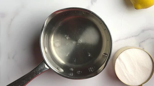 Small saucepan of clear syrup on a marble counter beside sugar and lemon close view of stovetop preparation for homemade lemonade sweetener and citrus drink recipe in a bright kitchen