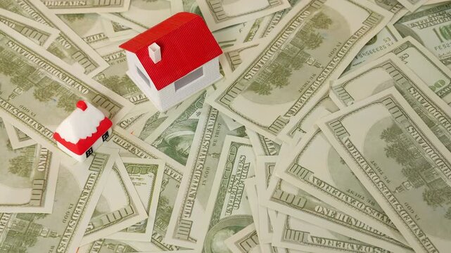 A small miniature model house placed on scattered stack of dollar bills banknotes represents real estate investment opportunities, home ownership, and the financial aspects of buying a property