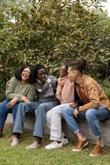 African American friends sitting on wooden bench at park, wearing sweaters and scarves, leaning in