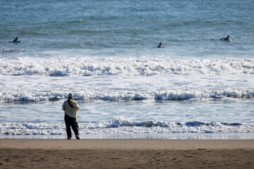 砂浜から沖のサーファーたちを眺める女性の後ろ姿