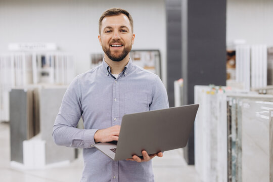 Man smiling, using a laptop in a showroom. He is a small business owner, sales manager, or client advisor