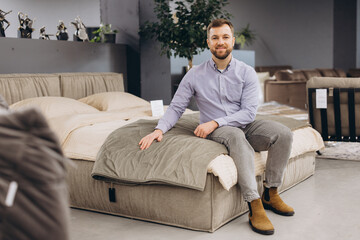 Obraz premium Happy man in a furniture showroom sitting on a comfortable bed, touching the bedding while shopping for home decor