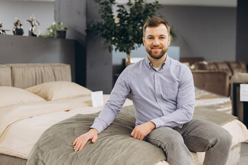 Fototapeta premium Smiling man sitting on a bed in a furniture store showroom, trying out mattresses, shopping for home interior items