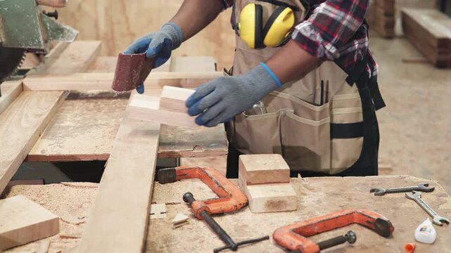 Woodworker in plaid shirt and apron shows a cut wood board inside a dusty carpentry workshop filled with tools and timber.