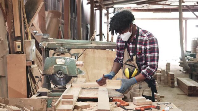 Woodworker in plaid shirt and apron shows a cut wood board inside a dusty carpentry workshop filled with tools and timber.