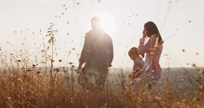 Family nature walk at sunset with children. Parents lead a kid across a golden meadow, enjoying summer light and gentle breeze. They move calmly on the ridge. Happy family together in nature.