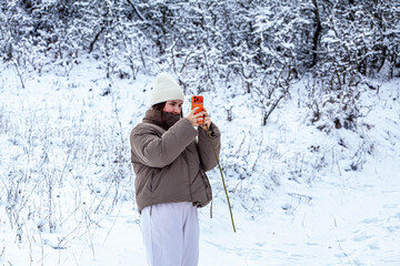 A very small and cute young lady surprised by the natural landscape of the winter season, posing on a very high-performance phone with snow on the branches of the trees.