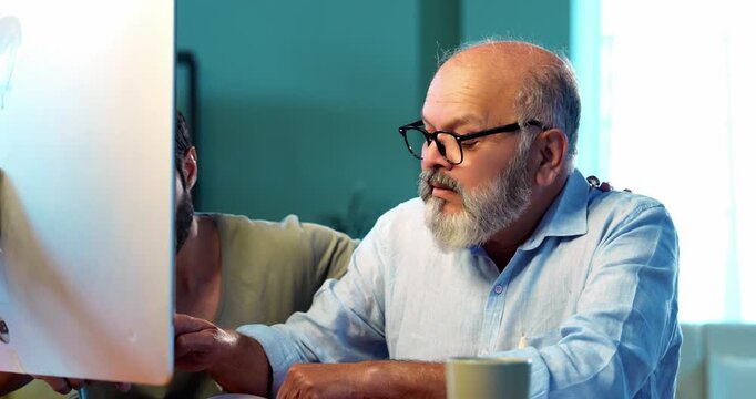 Indian young son teaching senior father how to use desktop computer at home in friendly way, elderly dad learning digital technology with smile while browsing internet together in modern living room