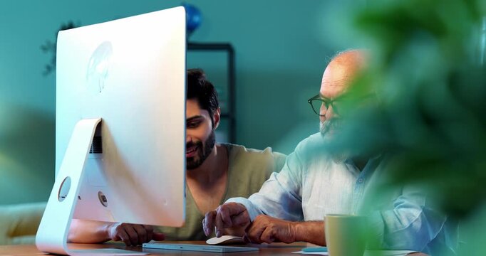 Indian young son teaching senior father how to use desktop computer at home in friendly way, elderly dad learning digital technology with smile while browsing internet together in modern living room