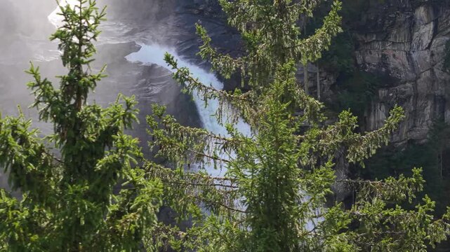 High-angle aerial footage of the Krimml Waterfalls in Austria during a sunny day. The video shows the powerful water flow crashing against rocks, surrounded by a lush pine forest and mist.