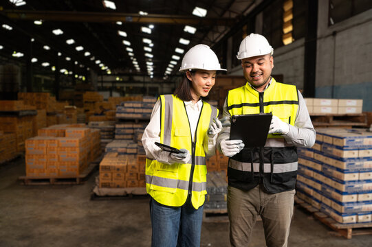 Two warehouse workers wearing safety gear discuss logistics while holding tablets, surrounded by stacks of boxes in the storage area.