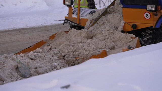 Orange municipal snowplow clearing dirty slush and ice from a city street after a heavy winter snowfall, pushing the frozen precipitation aside to ensure safe traffic flow and road accessibility