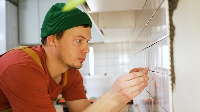 Concentrated male tiler in overalls meticulously installing white ceramic tiles on a kitchen wall, using plastic spacers to ensure even gaps for a professional home improvement project