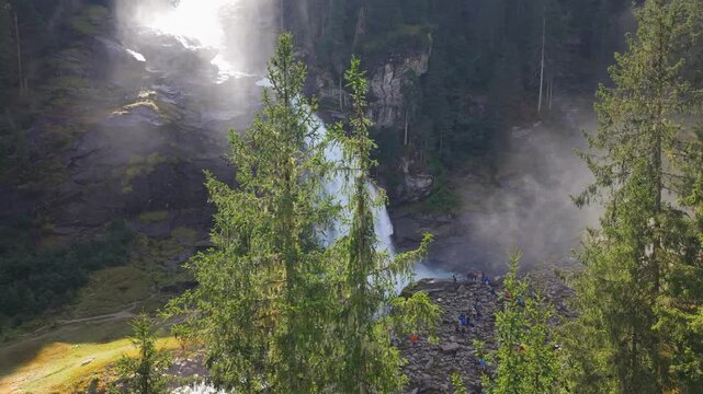High-angle aerial footage of the Krimml Waterfalls in Austria during a sunny day. The video shows the powerful water flow crashing against rocks, surrounded by a lush pine forest and mist.