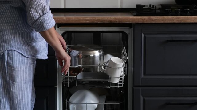 Woman in pajamas unloads a dishwasher and takes out clean kitchenware including pot, plates, cups, and glass bowls. Rear view with cropped frame showing hands, shoulders, and waist only.