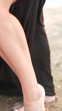 Barefoot, black dress, woman showing leg on sandy ground. Vertical video.