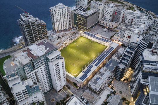 Aerial view of a brightly lit football field nestled among modern buildings near the coast, contrasting with the deepening twilight, Sliema, Sliema, Malta.