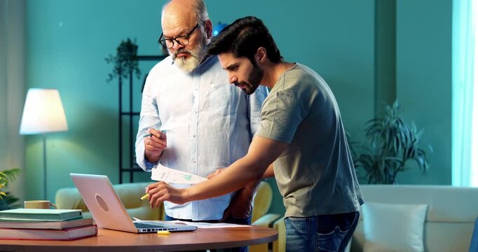 Indian young son and senior father filing online tax return together using laptop while standing at table in modern home, discussing financial reports, income statements yearly accounting documents